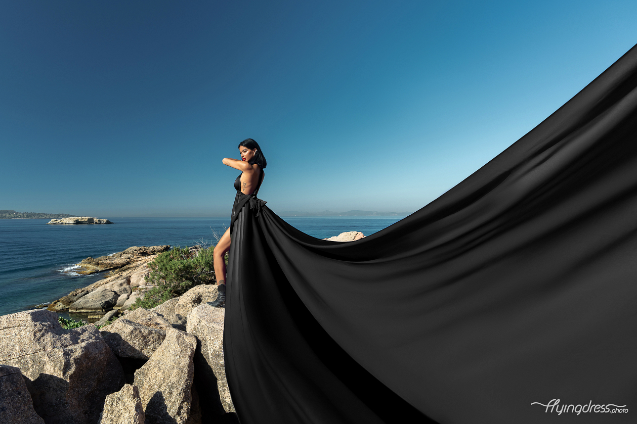 A woman in a flowing black dress stands on rocky cliffs with a vast blue sea and distant islands in Kavouri, Athens, creating a striking contrast against the bright sky.